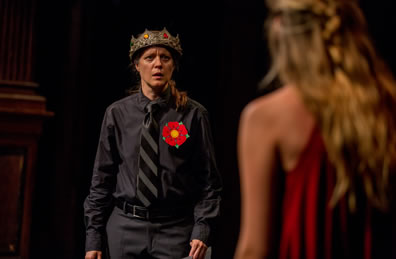 Henry in black shirt and pants with black and gray wide-striped tie and a red rose on his shirt and a crown on his head looks with concern on an unidentified  woman, back to the camera.